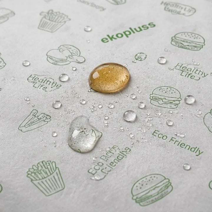 A close-up of a textured white surface printed with green illustrations of fast food items and words like 'ekopluss' and 'Eco Friendly', with two water droplets resting on top.