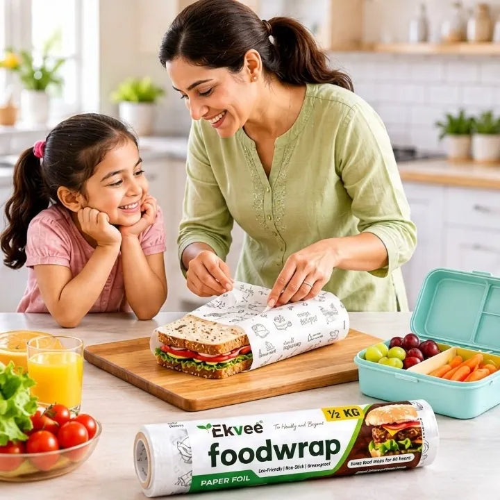 A woman and a young girl in a bright kitchen setting, with the woman wrapping a sandwich in Ekvee paper food wrap placed on a wooden cutting board. A roll of Ekvee food wrap is shown in the foreground, with fresh vegetables, juice, and a lunchbox nearby.